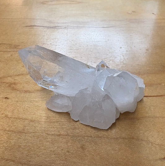 A raw quartz crystal cluster with a prominent pointed crystal terminus resting on a light brown wooden surface.