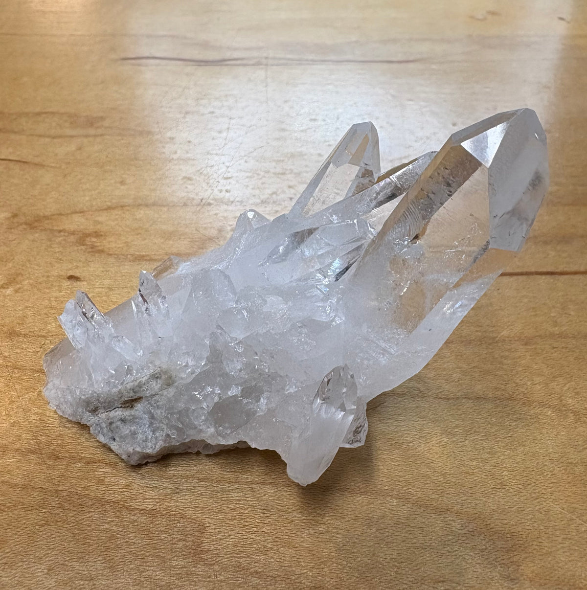 A natural clear quartz crystal cluster with multiple pointed tips resting on a light brown wooden surface.