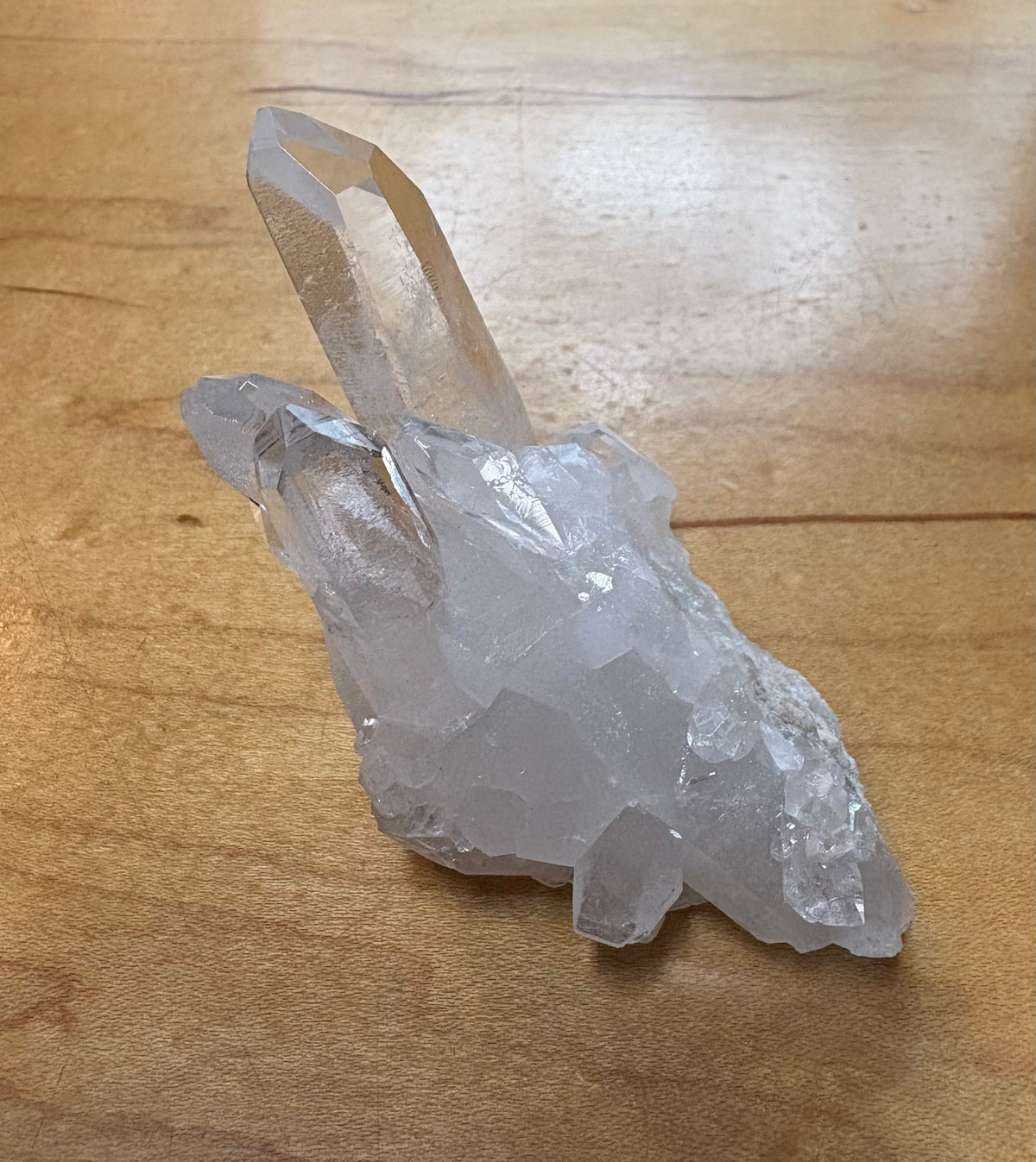 A raw clear quartz crystal cluster with a prominent tall point, resting on a light brown wooden surface.