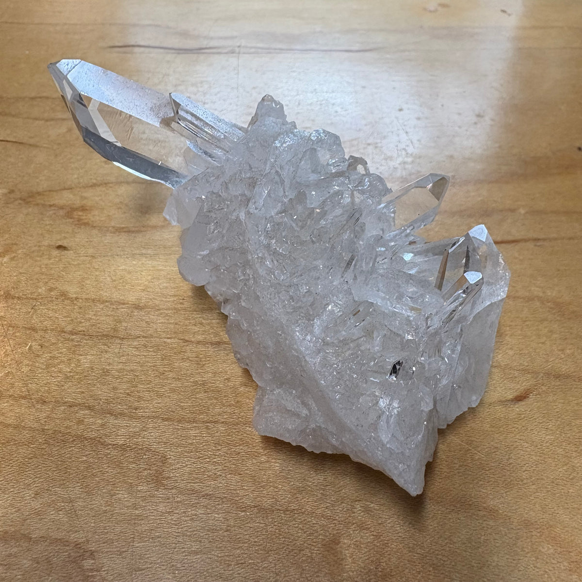 A raw, rough crystal cluster with multiple clear, faceted points resting on a light brown wooden surface.