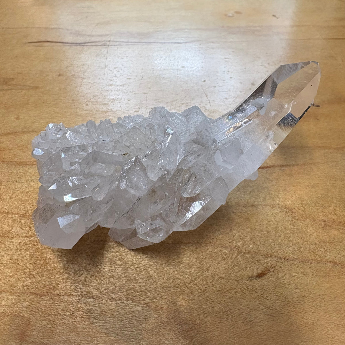 A natural clear quartz crystal cluster with a prominent pointed crystal terminus, resting on a light brown wooden surface.