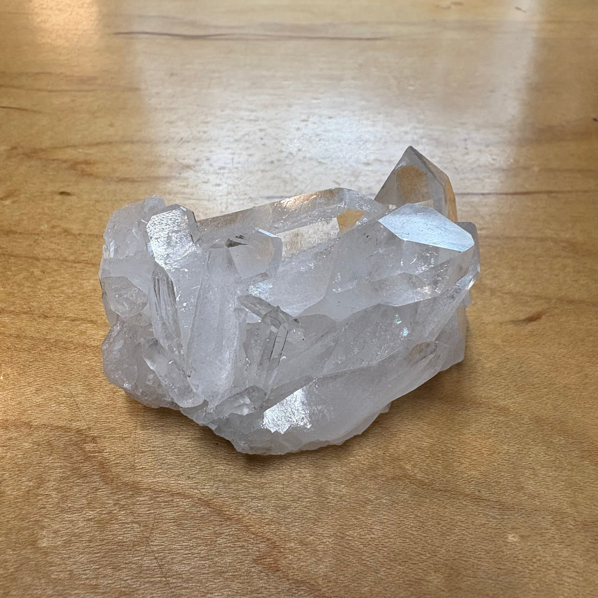 A raw, unpolished cluster of clear quartz crystals with sharp geometric facets, resting on a light brown wooden surface.