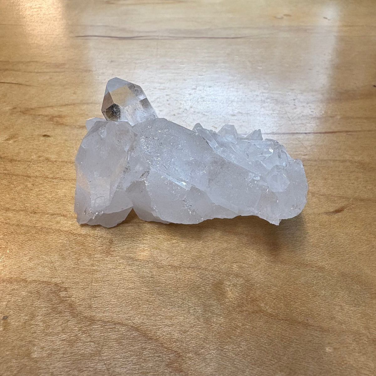 A raw clear quartz crystal cluster with multiple natural points and rough faceted edges, resting on a light brown wooden tabletop.