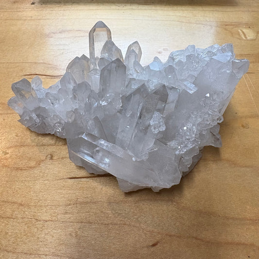 A natural clear quartz crystal cluster with multiple pointed and faceted quartz tips, displayed on a light brown wooden surface.