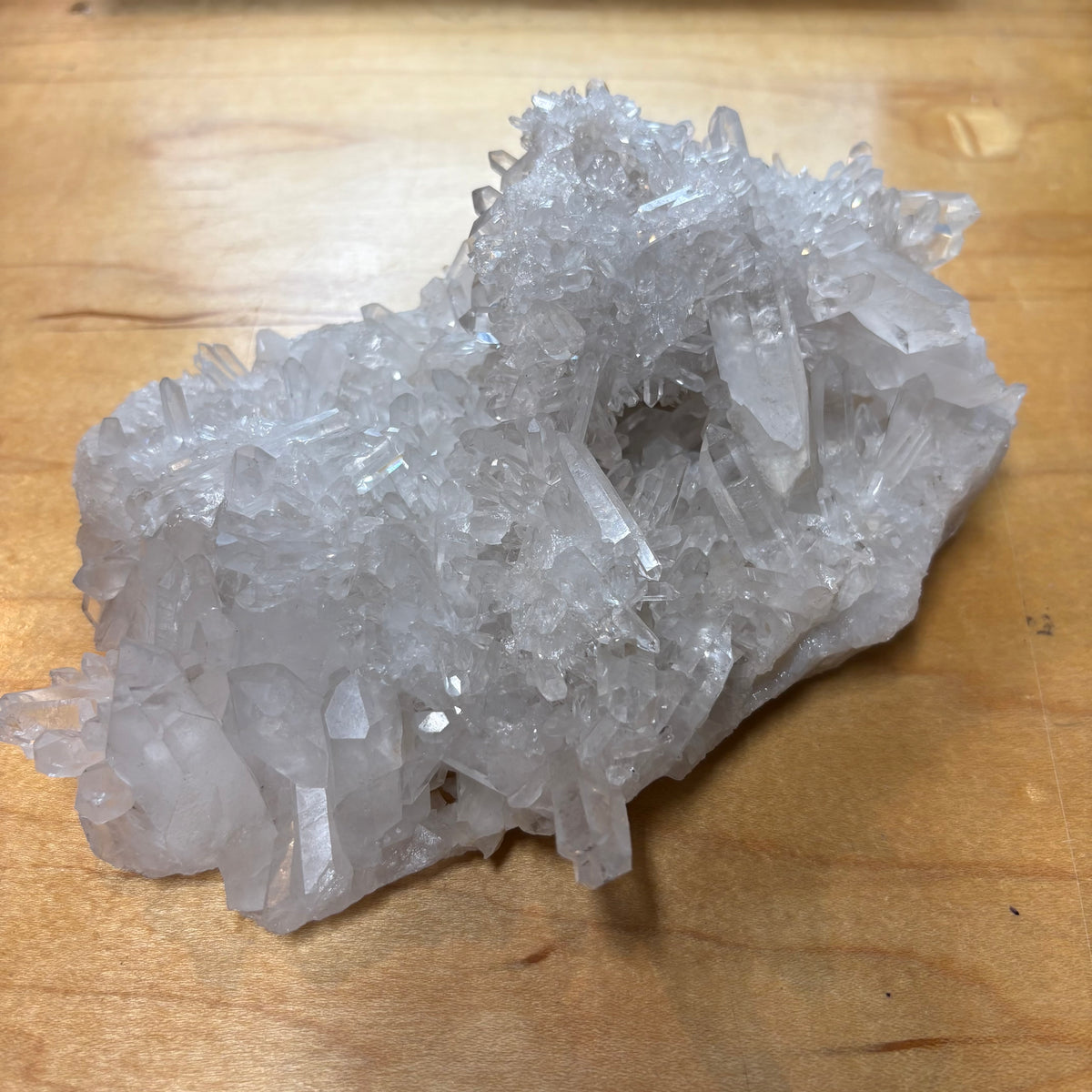 A natural clear quartz crystal cluster with multiple sharp, branching points, resting on a light brown wooden surface.