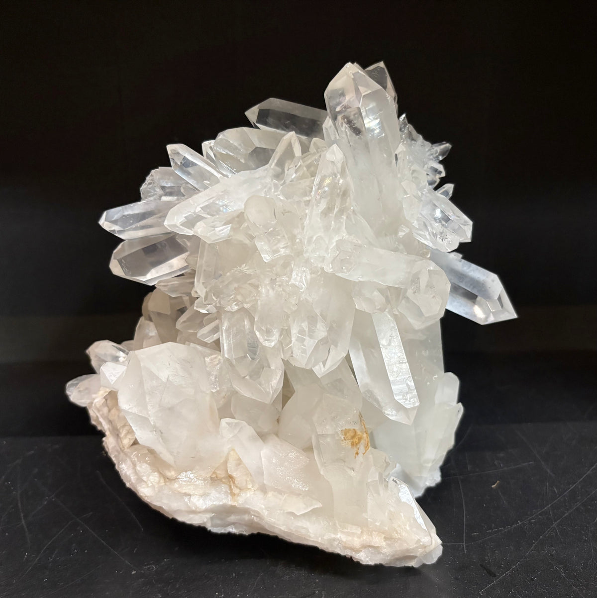 A natural quartz crystal cluster with clear, faceted quartz points rising from an off-white matrix, displayed on a dark black background.