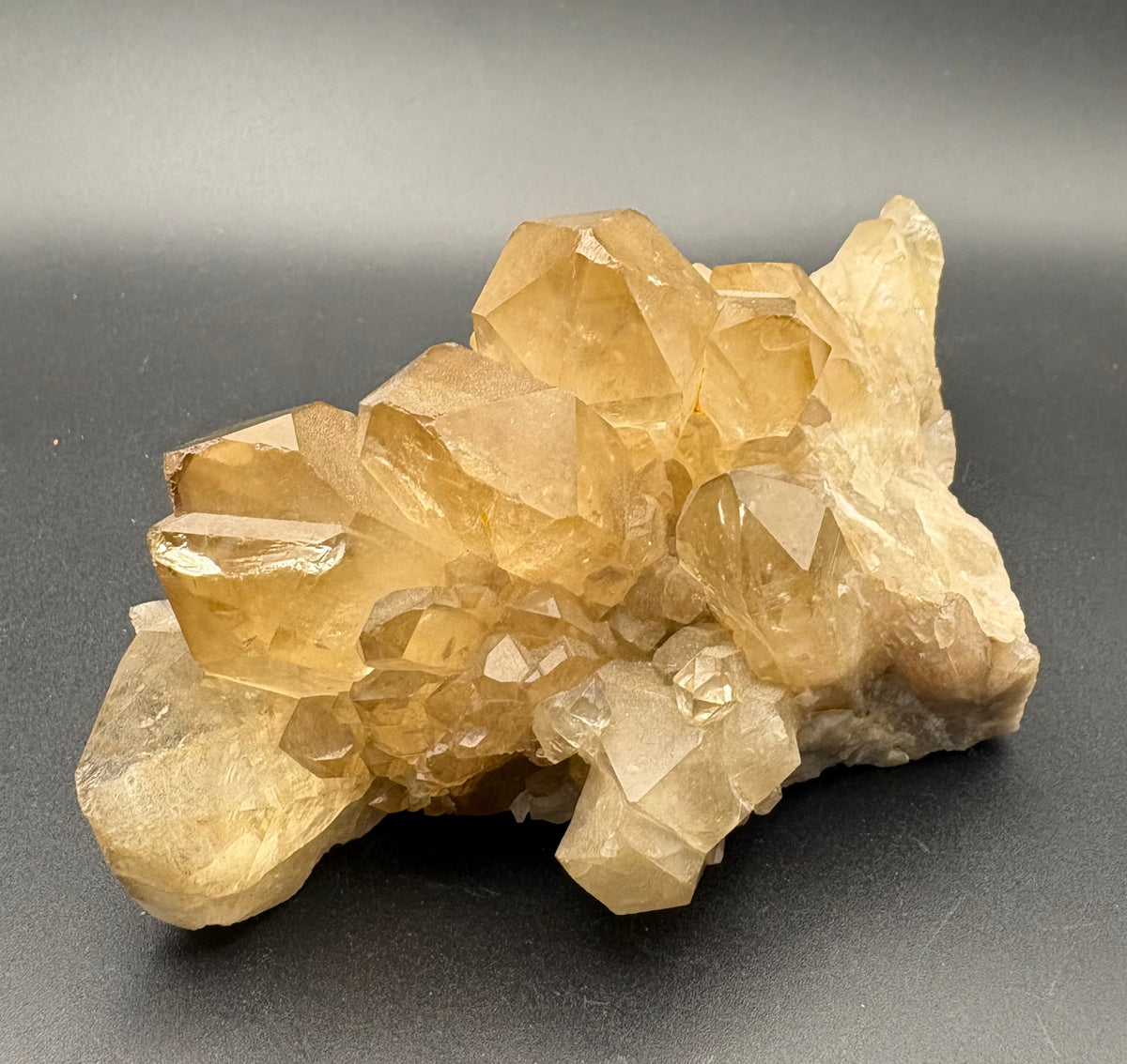 A natural quartz crystal cluster specimen with translucent golden-yellow to light brown facets, displayed on a dark gray background.