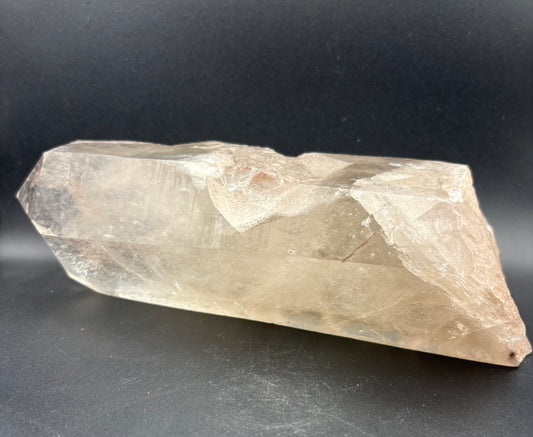 A raw, smokey quartz crystal point with a warm, translucent beige-tan hue, displayed on a dark, neutral background.