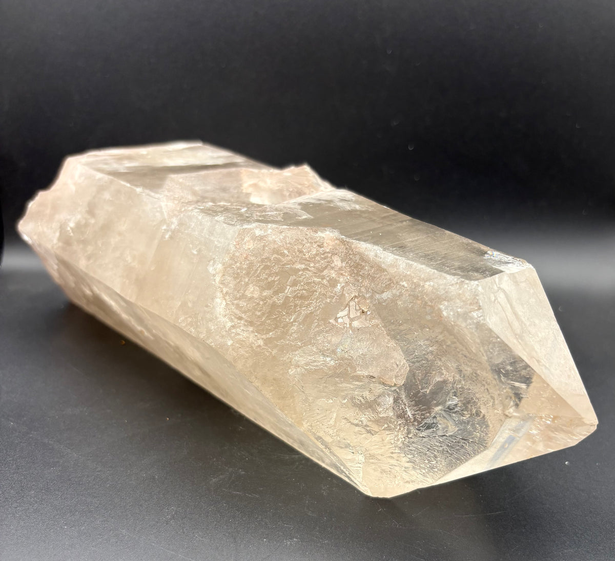 A raw, smoky quartz crystal point with a warm, translucent beige-tan hue, displayed on a dark, neutral background.