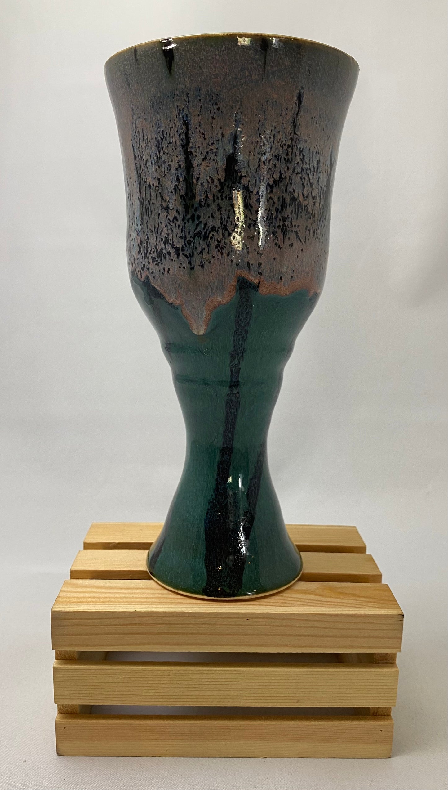 Black, green, lavender  ceramic chalice on a wooden stand against a white background