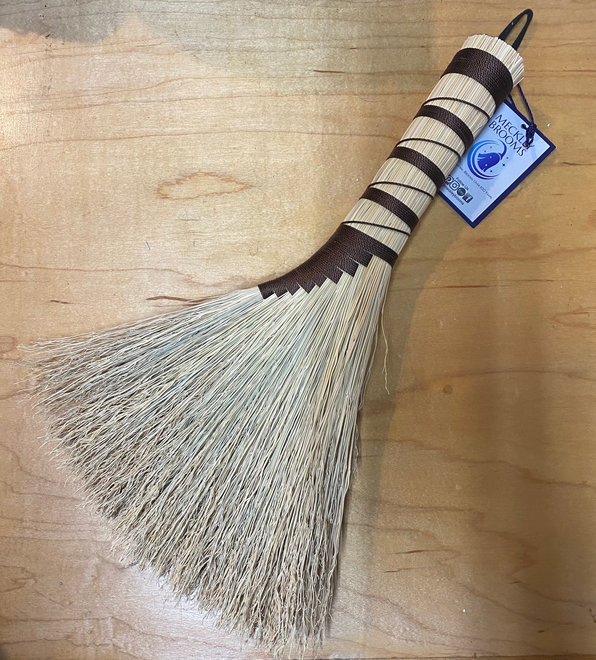 Broom with a wooden handle and straw bristles on a wooden surface, featuring a brand label.