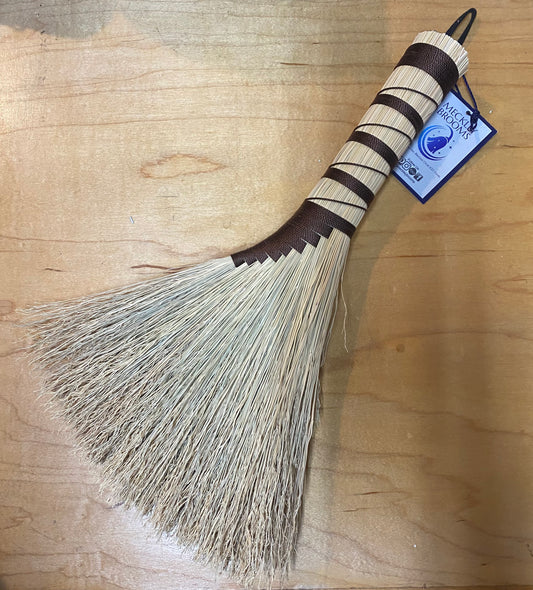 Broom with a wooden handle and straw bristles on a wooden surface, featuring a brand label.