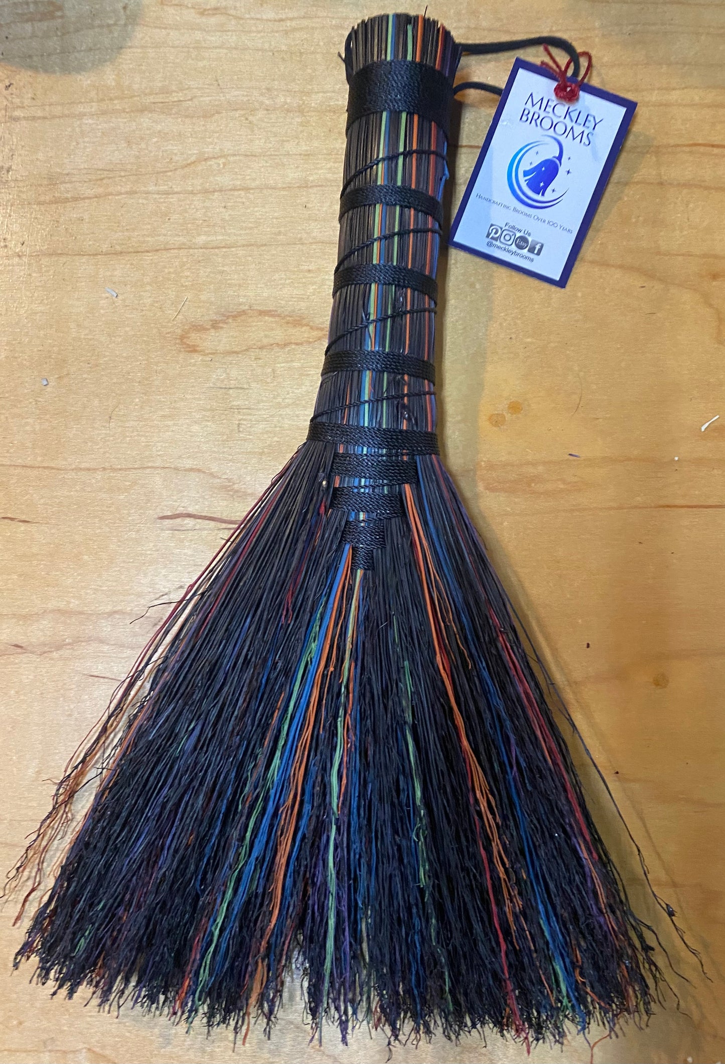 Decorative broom with multicolored bristles on a wooden surface, featuring a tag.