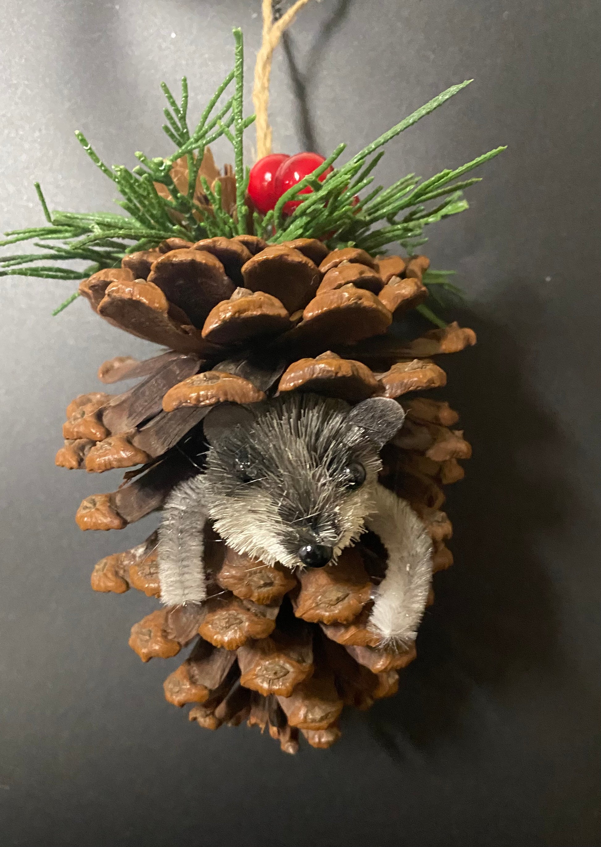 Decorative ornament with a pine cone, small animal figure, and greenery on a dark background