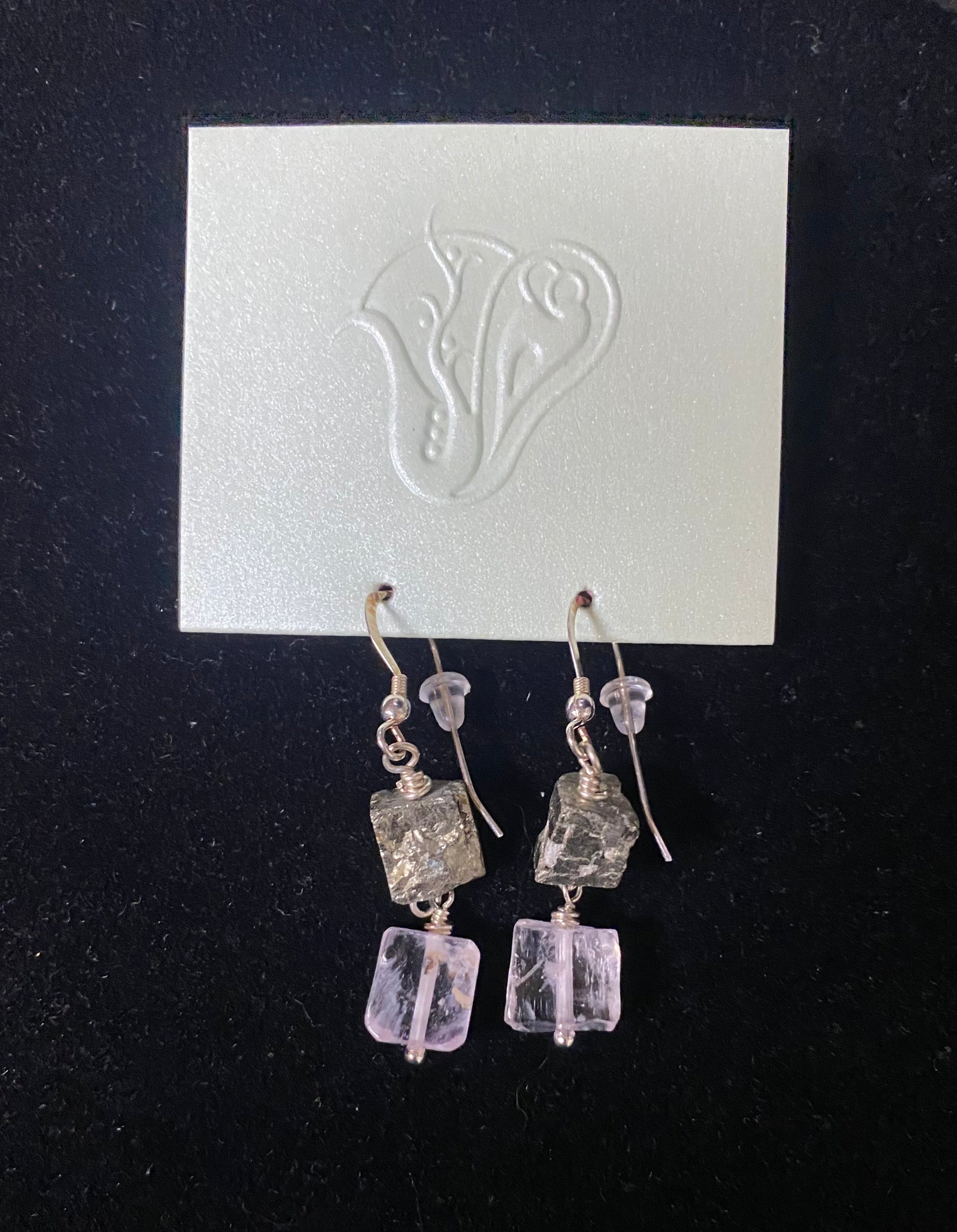 Pair of earrings with square and rectangular stones on a black background with a white card featuring a design.