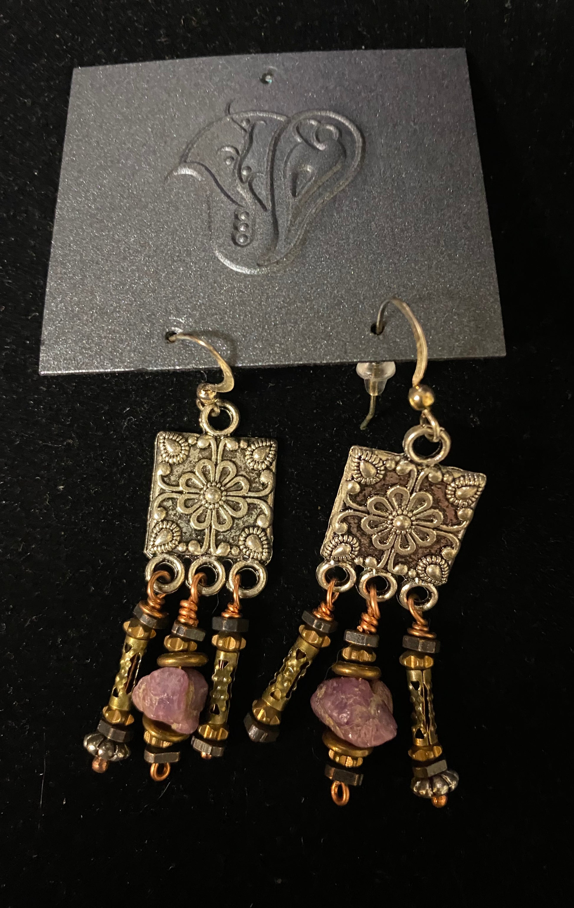 Pair of ornate earrings with purple stones on a black card with a brand logo.