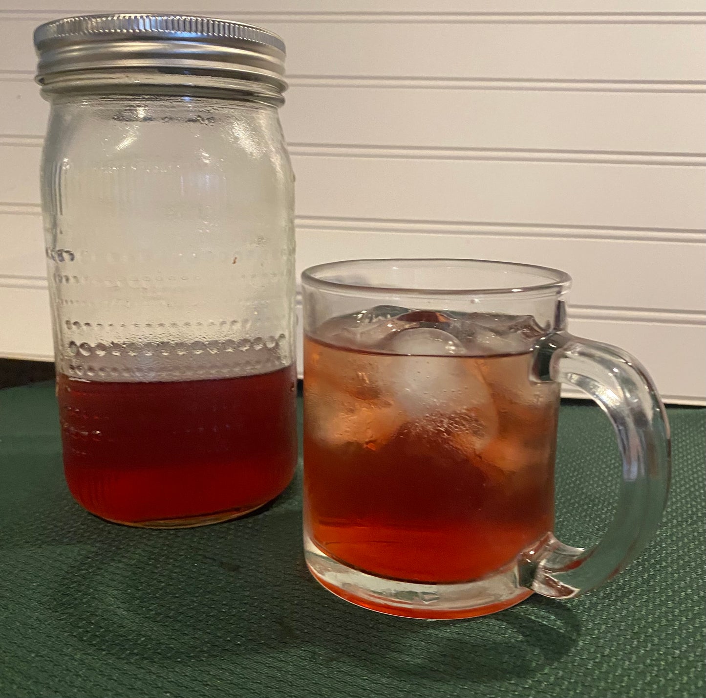 Mason jar and a glass with red colored iced herbal Tea with ice cubes on a green table with a white background
