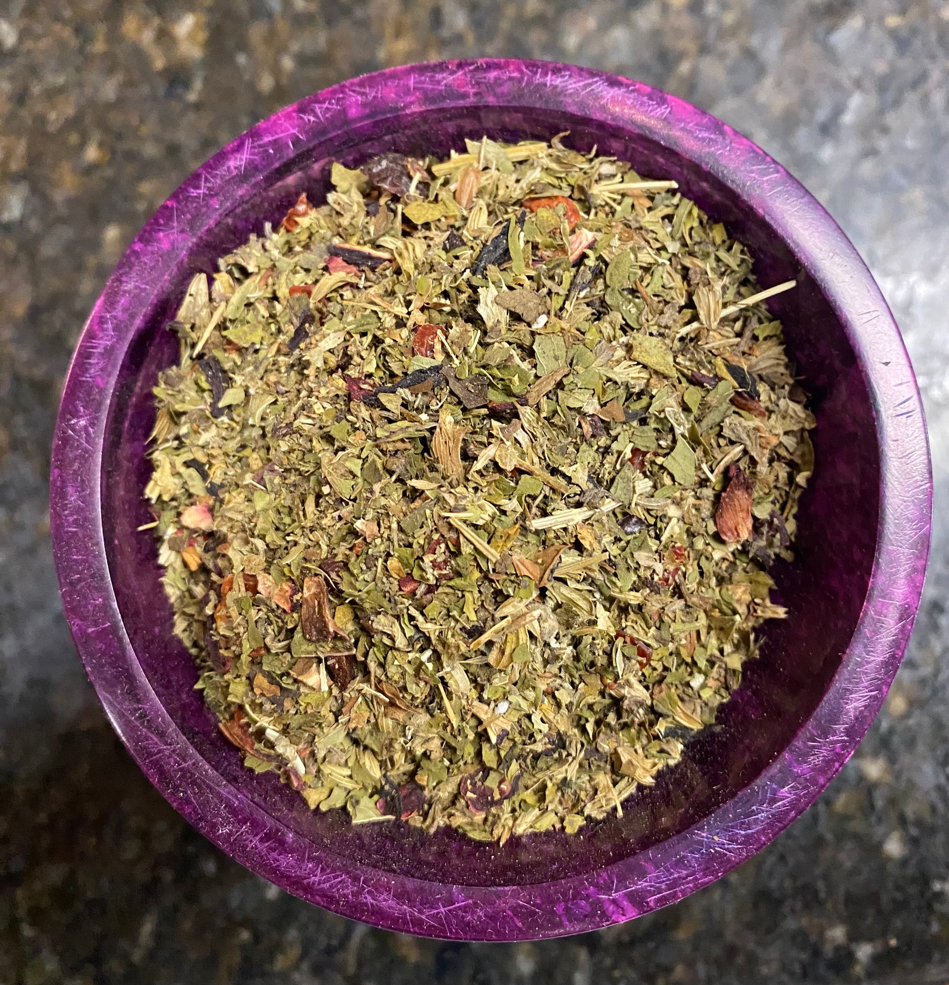 A top-down view of dried, chopped green herbs or tea leaves piled inside a round purple wooden bowl, set on a dark, mottled stone surface.