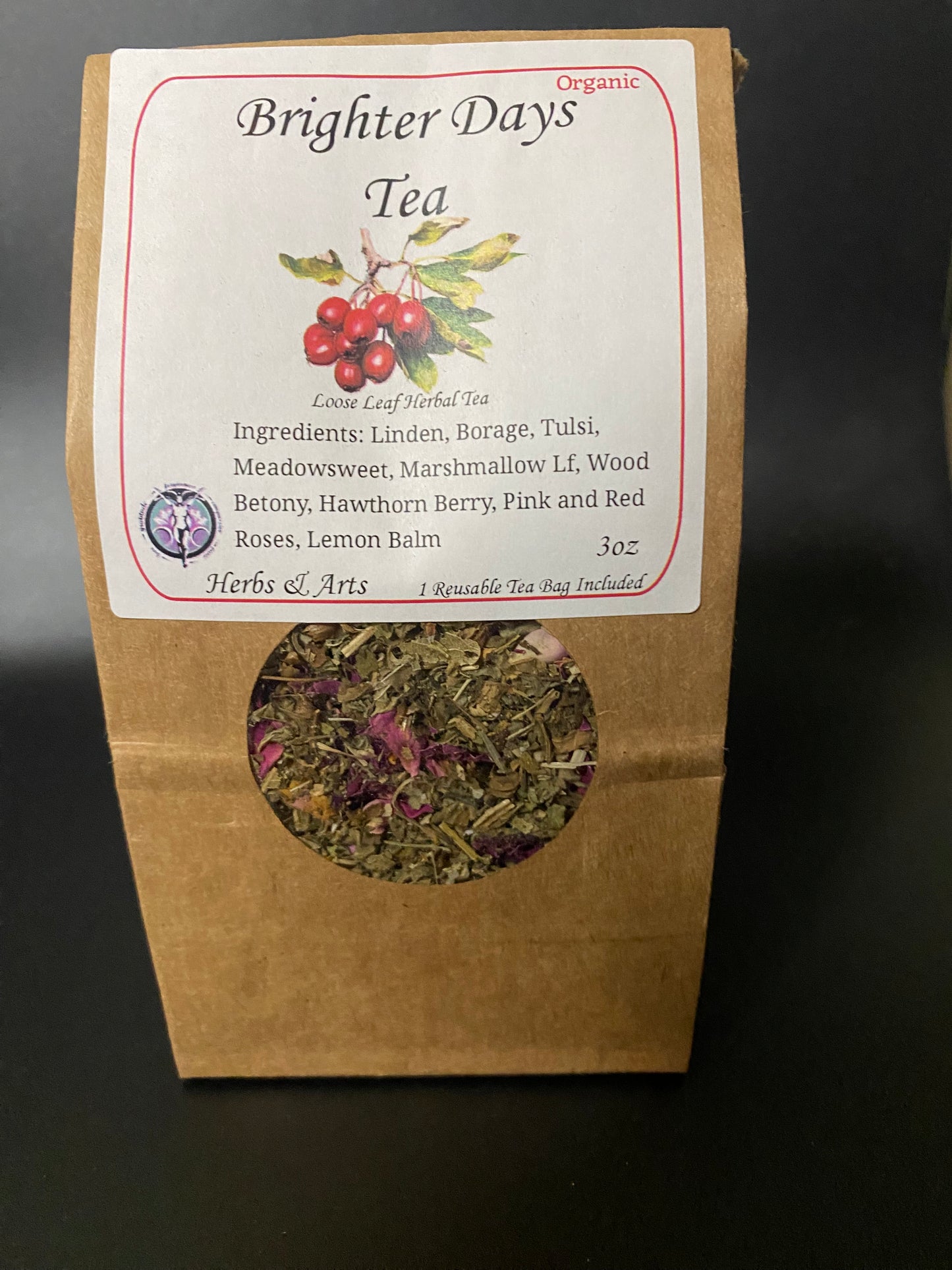 Brighter Days Organic Tea in a brown kraft paper pouch with a white label and a circular cutout showing dried tea leaves and rose petals, photographed on a dark background.