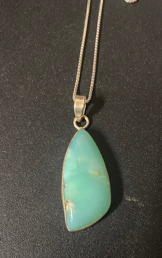 A silver chain necklace with a teardrop-shaped light green stone pendant, displayed on a dark textured surface.