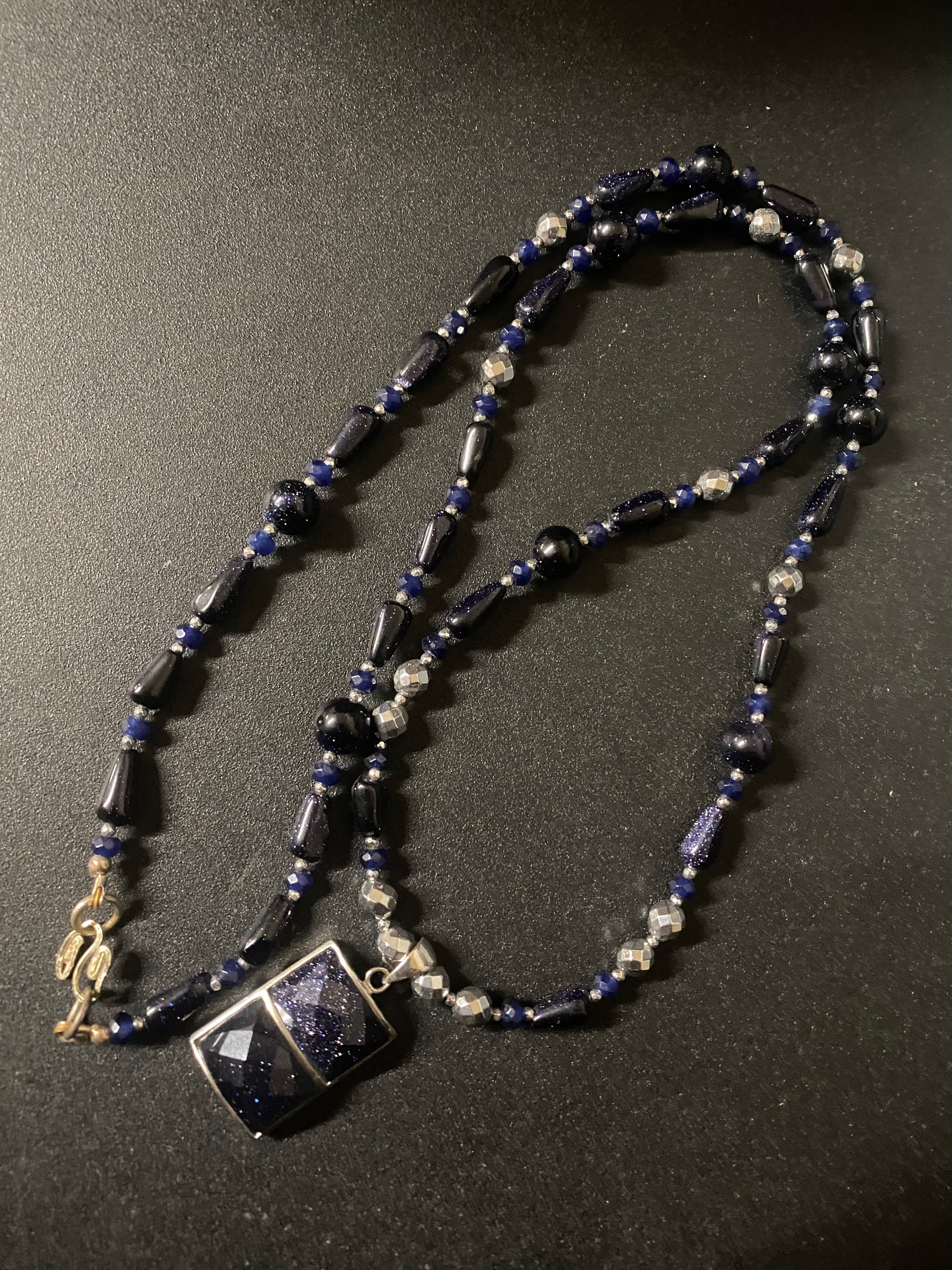 A beaded necklace featuring dark blue goldstone, blue faceted beads, and silver-toned spacer beads, with a square dark blue pendant set in silver, displayed on a dark textured background.
