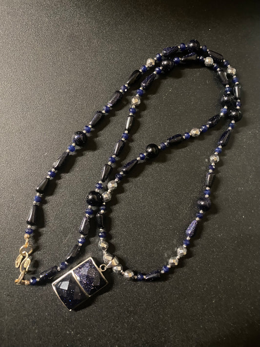 A beaded necklace featuring dark blue goldstone, blue faceted beads, and silver-toned spacer beads, with a square dark blue pendant set in silver, displayed on a dark textured background.