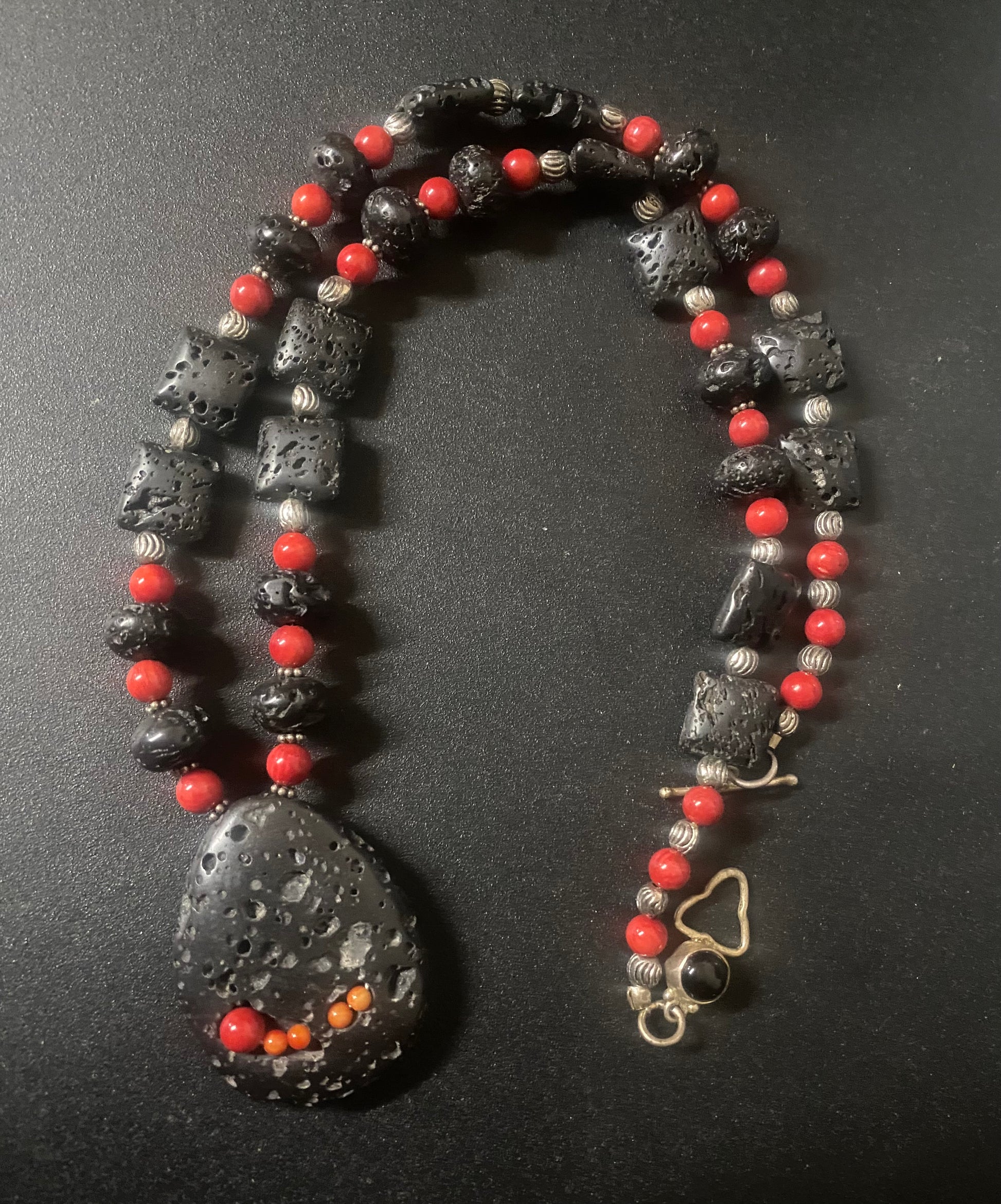 A beaded necklace featuring large black porous lava stones, small red beads, and silver-toned metallic beads, with a large teardrop-shaped black stone pendant and a gold-tone clasp on a dark background.