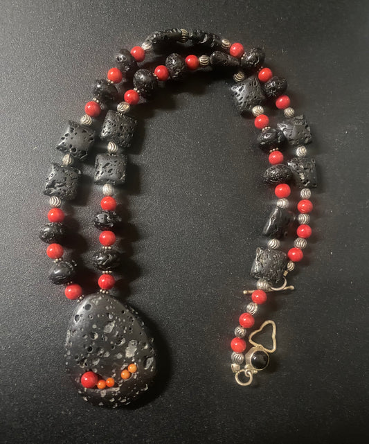 A beaded necklace featuring large black porous lava stones, small red beads, and silver-toned metallic beads, with a large teardrop-shaped black stone pendant and a gold-tone clasp on a dark background.