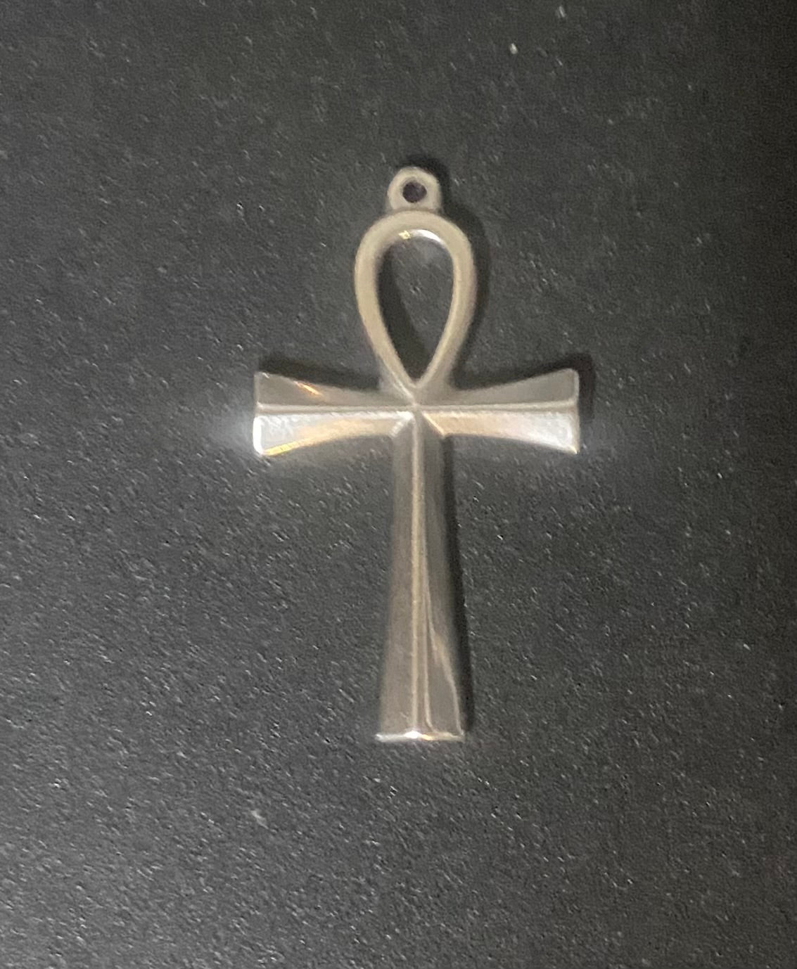A polished silver-toned metal ankh pendant with a flat, geometric design, shown on a dark textured background.