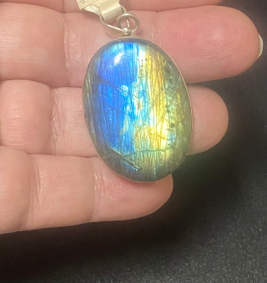 A silver pendant with an oval labradorite stone held in a person's hand. The stone displays vibrant blue, green, and yellow iridescent flashes.