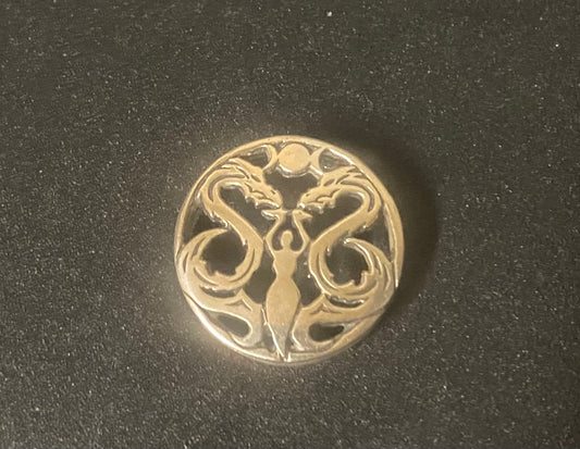 A round silver-toned metal pendant featuring an intricate cutout design of two stylized figures facing each other, set against a dark textured background.