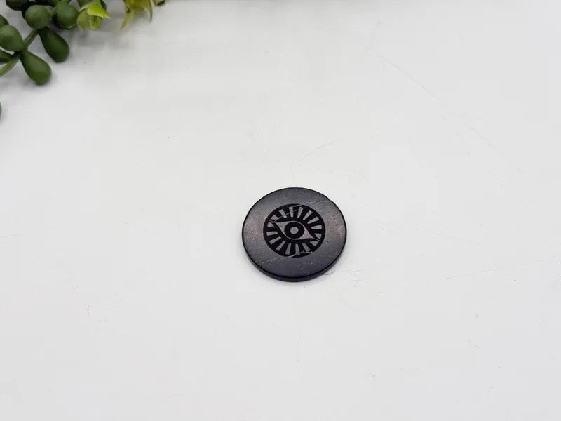 A single round black button with a sunburst or wheel-like cutout pattern in the center, placed on a plain white surface with a small green plant visible in the top-left corner.