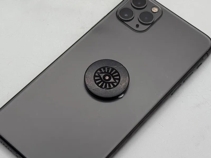 A close-up of a black smartphone back with a circular black tile featuring an eye-of-providence symbol, placed on a light gray surface.