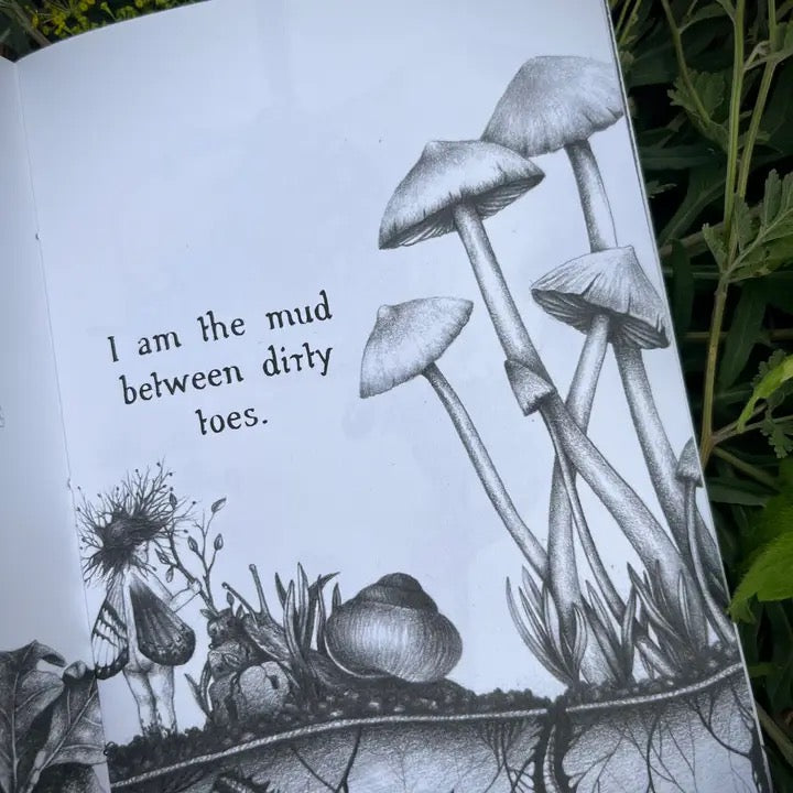 An open book or notebook page with black-and-white illustrated mushrooms, a small fairy-like figure, and the text, 'I am the mud between dirty toes.' photographed outdoors with green leaves visible around the edges.