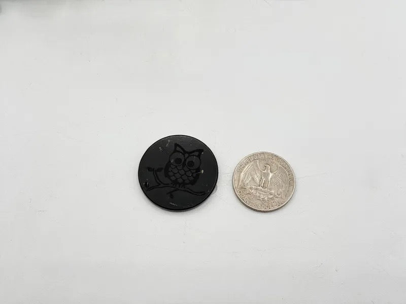 A small black oval button with an owl design next to a US quarter coin for scale, shown on a plain white background.