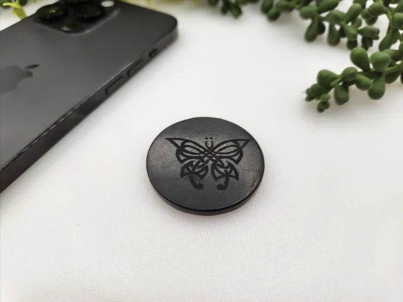 A round black magnetic phone accessory with an engraved butterfly design, shown on a white surface next to a black smartphone and green plant leaves.