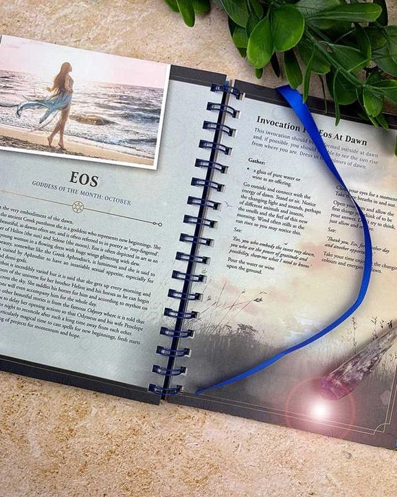 An open spiral-bound journal or prayer book laid on a beige stone surface, with printed text pages visible, a small beach photo insert on the left page, and a bright blue ribbon draped across the right page. Green leaves and a small crystal pendant are placed around it as styled props.