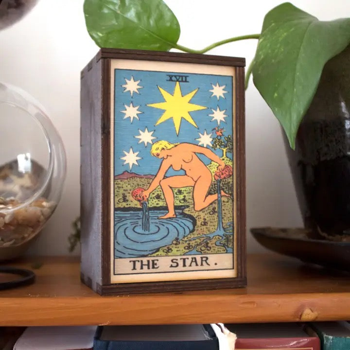 Wooden box with 'The Star' tarot card design on a shelf with plants in the background