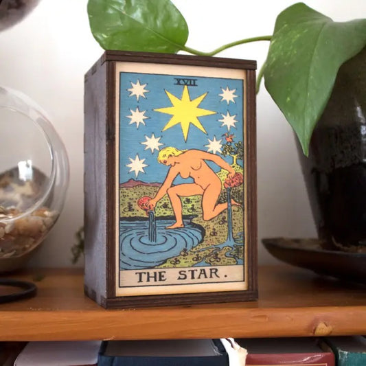 Wooden box with 'The Star' tarot card design on a shelf with plants in the background
