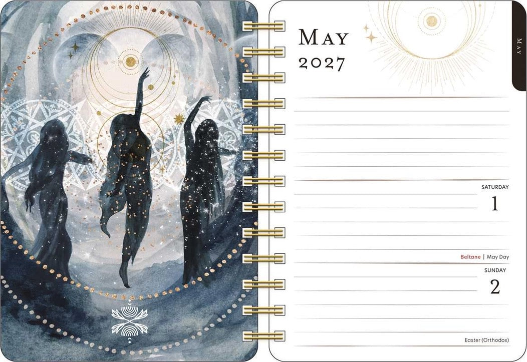 A spiral-bound May 2027 calendar page with a mystical illustrated cover showing three dark silhouetted figures against a blue-gray celestial background with gold circular symbols. The right page shows lined date slots for Saturday and Sunday, May 1 and 2.