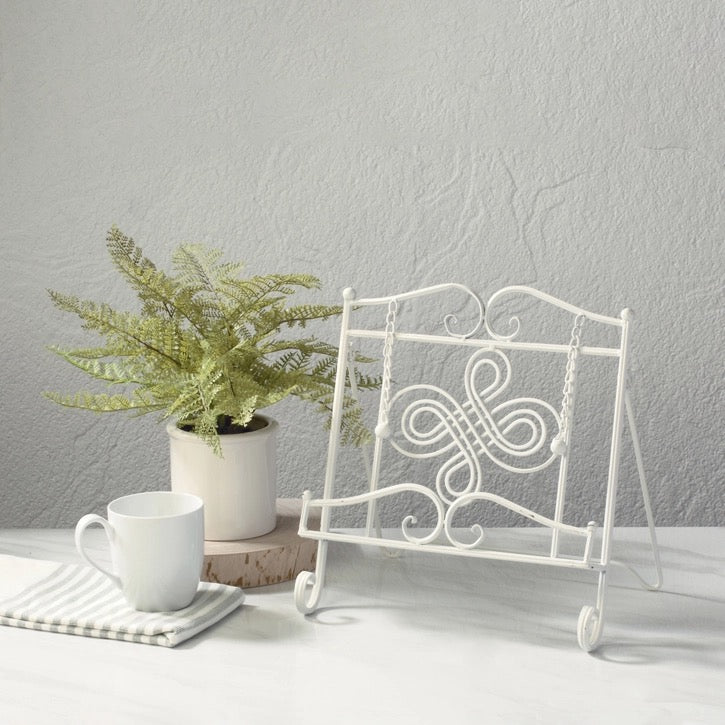 A white metal decorative display stand with scrollwork and hanging rings, styled on a tabletop beside a potted fern, a white mug, and a striped napkin against a light gray textured wall.
