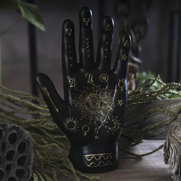 A black hand-shaped decorative object with gold occult and astrological symbols, including a central eye motif and zodiac signs, displayed on a rustic surface with dried greenery and dark background elements.