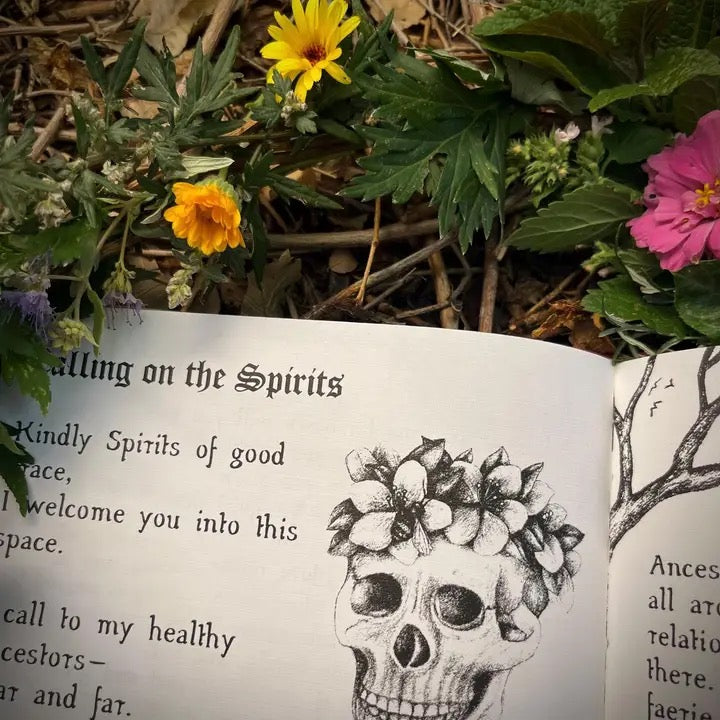 An open book spread with gothic text reading 'Calling on the Spirits' and a black-and-white skull illustration with flowers in its teeth, photographed outdoors among green leaves and small yellow and pink flowers.