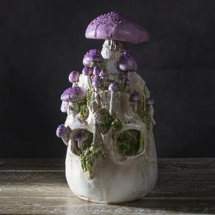 A decorative skull sculpture with embedded purple mushrooms, green moss, and a textured white bone-like body, displayed on a rustic wooden surface against a dark background.