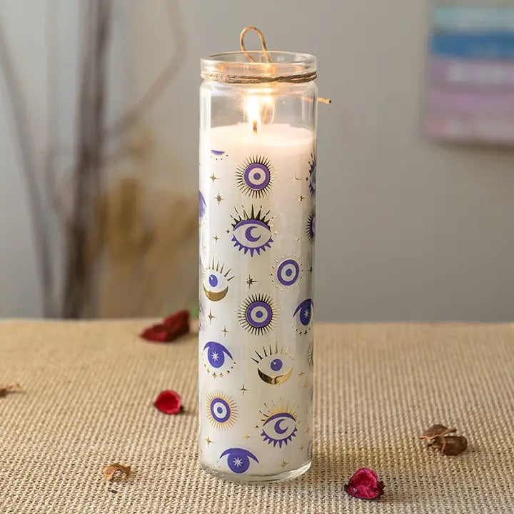 A tall clear glass candle holder filled with white wax and decorated with blue, purple, and gold eye and moon motifs, lit with a warm flame. The candle sits on a beige textured surface with scattered rose petals and dried botanicals in the background.