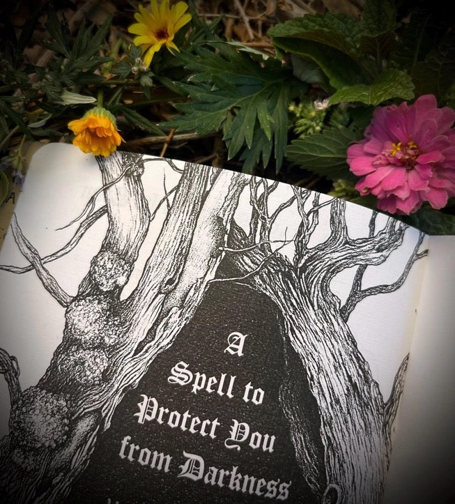 An open book or card with black-and-white illustrated tree branches and the text 'A Spell to Protect You from Darkness,' styled with flowers and green leaves around it.