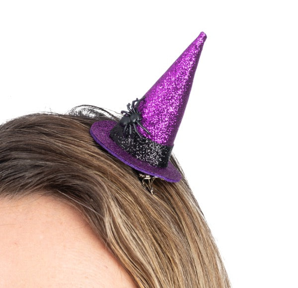 A purple glitter witch hat hair clip with a black band and spider decoration, shown clipped to brown hair against a white background.