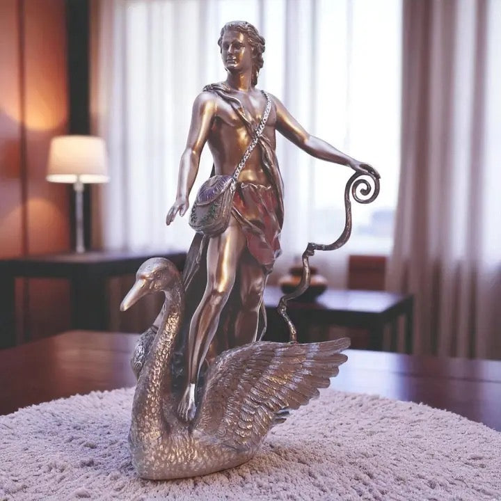 A bronze-finished sculpture of a classical male figure riding a swan, displayed on a textured light gray rug in a softly lit indoor room with curtains and furniture in the background.