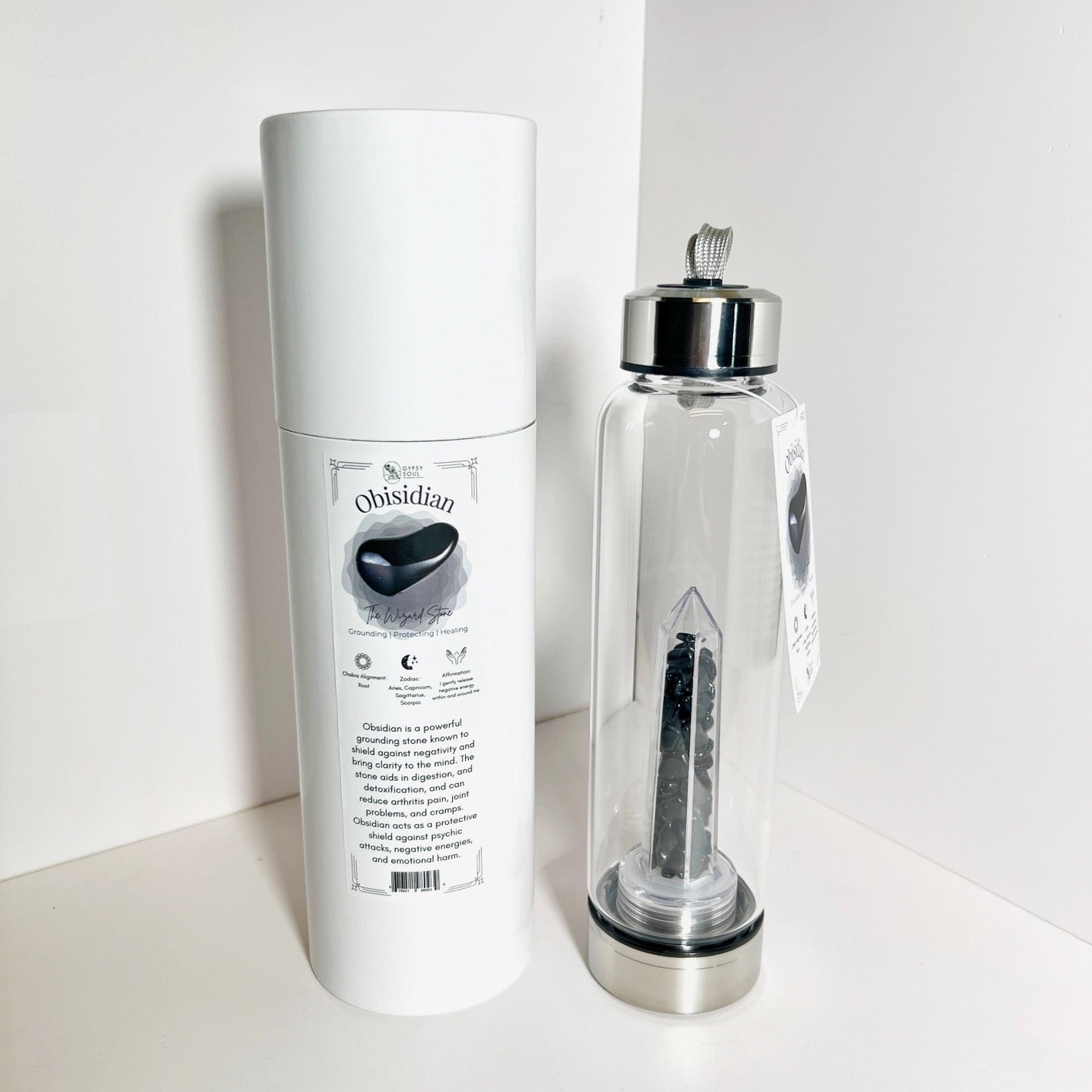 Glass bottle with silver cap and black stone, next to a white cylindrical packaging on a white background