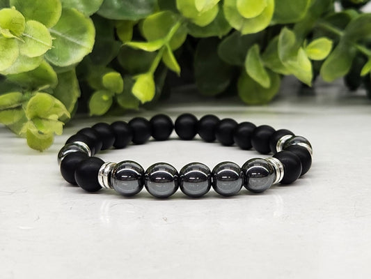 Black and gray beaded bracelet on a white surface with green leaves in the background